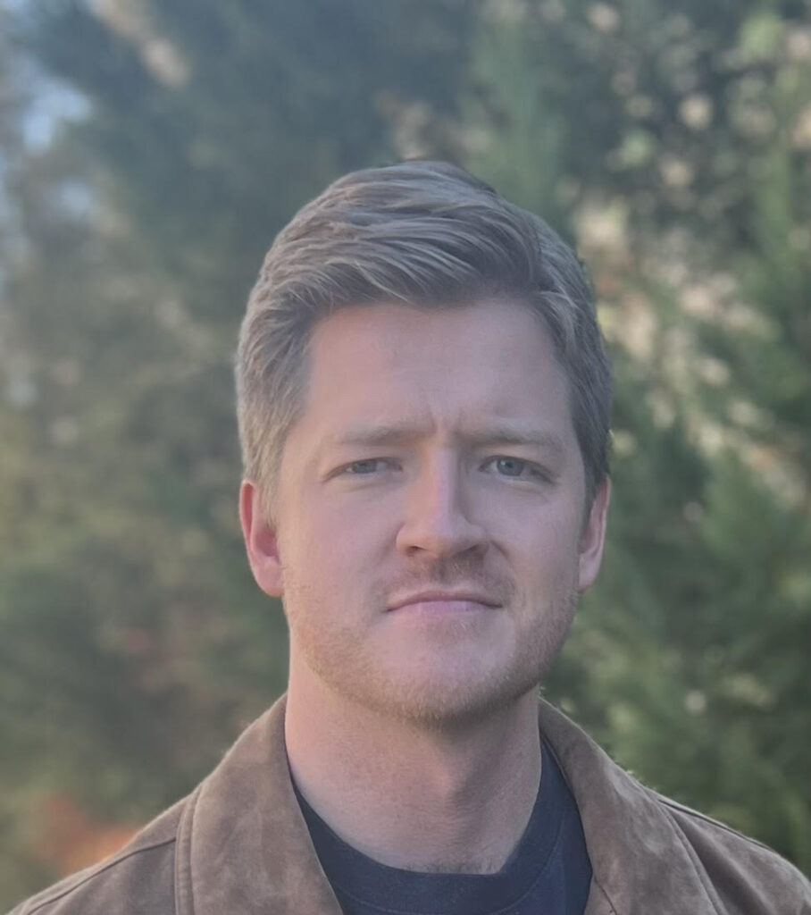 A man with short, light brown hair and light stubble stands outdoors with a serious expression. He is wearing a brown suede jacket over a black shirt. The background consists of blurred green trees, suggesting a natural setting.