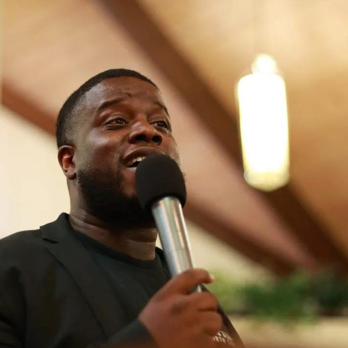 A man speaking into a microphone inside a building with wooden ceiling beams and warm lighting. He is wearing a dark blazer and a black shirt, and appears to be addressing an audience.