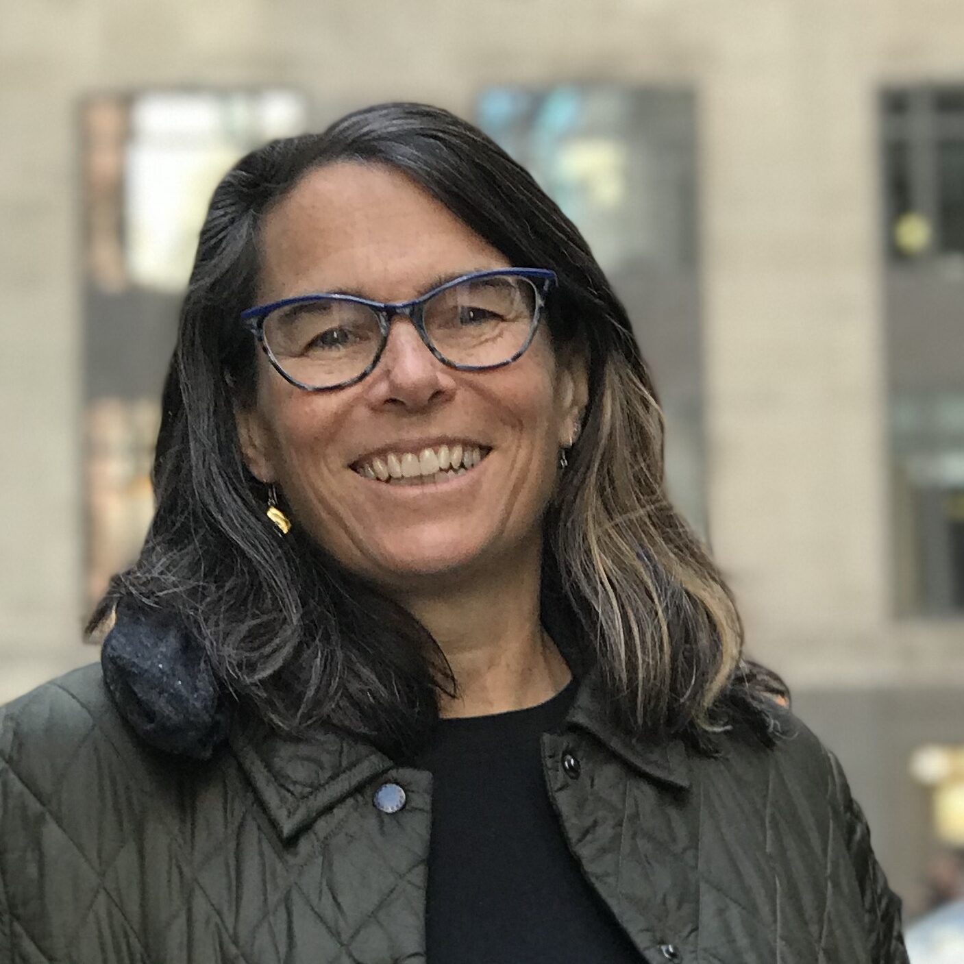 Rose Hoban, a woman with shoulder-length dark hair, accented with a blonde streak, smiles at the camera while standing outdoors in an urban setting. She wears blue-framed glasses, gold earrings, a black shirt, and a quilted olive-green jacket. The blurred background features a modern building with large windows reflecting light.