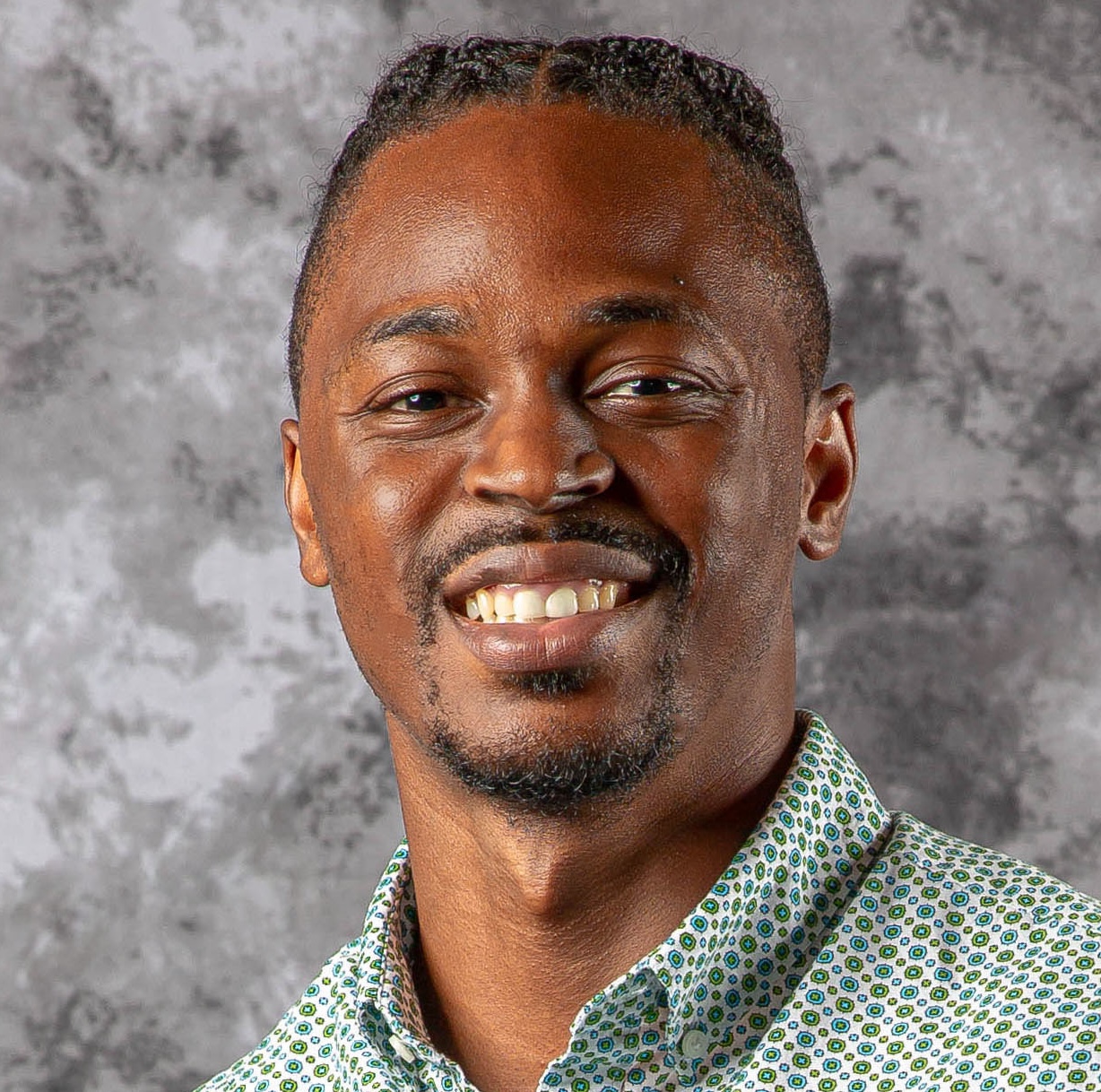 Martin Henson, a smiling man with braided hair, wearing a patterned button-up shirt, posed in front of a gray marbled background.