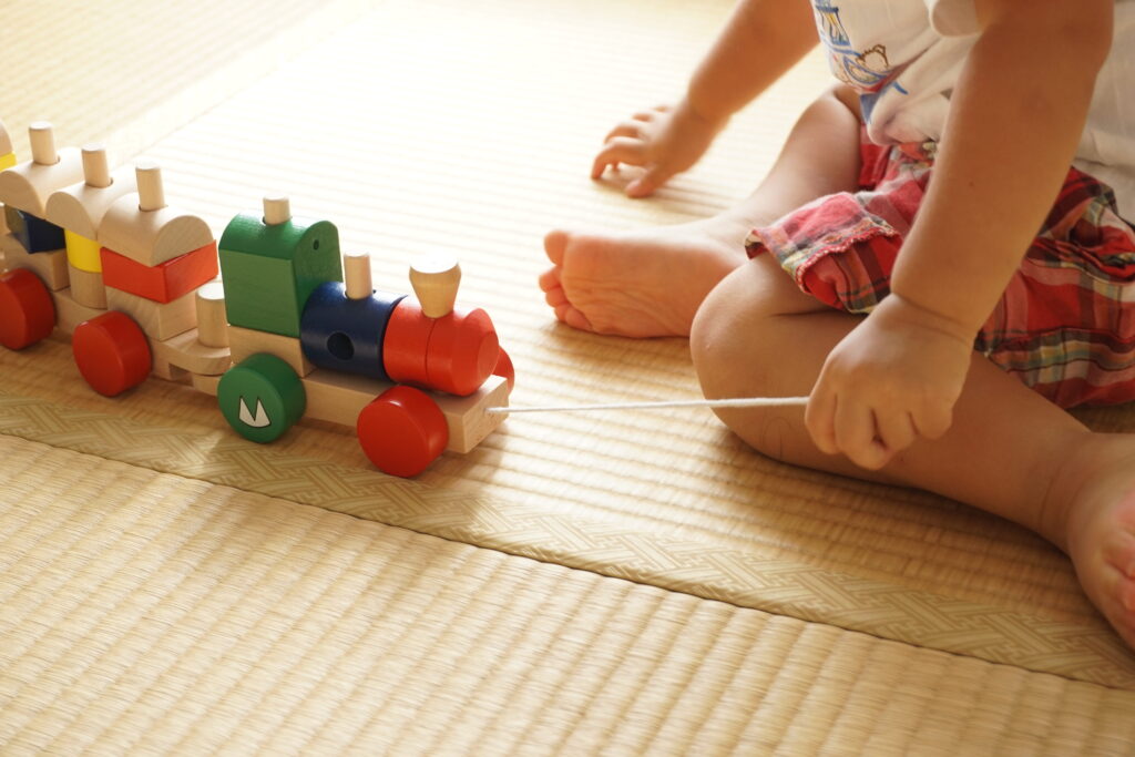 A toddler sits on a woven mat floor, playing with a colorful wooden toy train. The child, dressed in a white shirt and red plaid shorts, is holding a string attached to the train, pulling it gently. Only the child's arms, legs, and part of their outfit are visible.