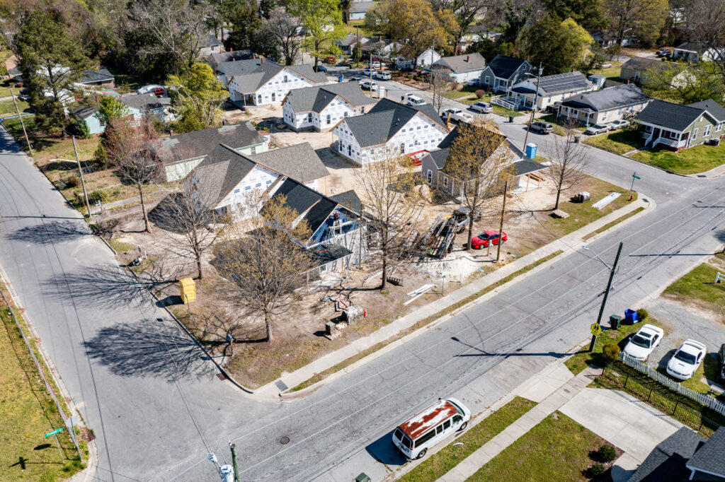 A bird's-eye view of a residential neighborhood under construction, with multiple houses in various stages of completion. Most homes have black roofs, and some still have visible white weatherproofing wrap. Construction materials and equipment are scattered throughout the site. Leafless trees line the streets, and a red car and a white van are parked nearby. The neighborhood appears to be in a suburban or small-town setting with paved roads and sidewalks.