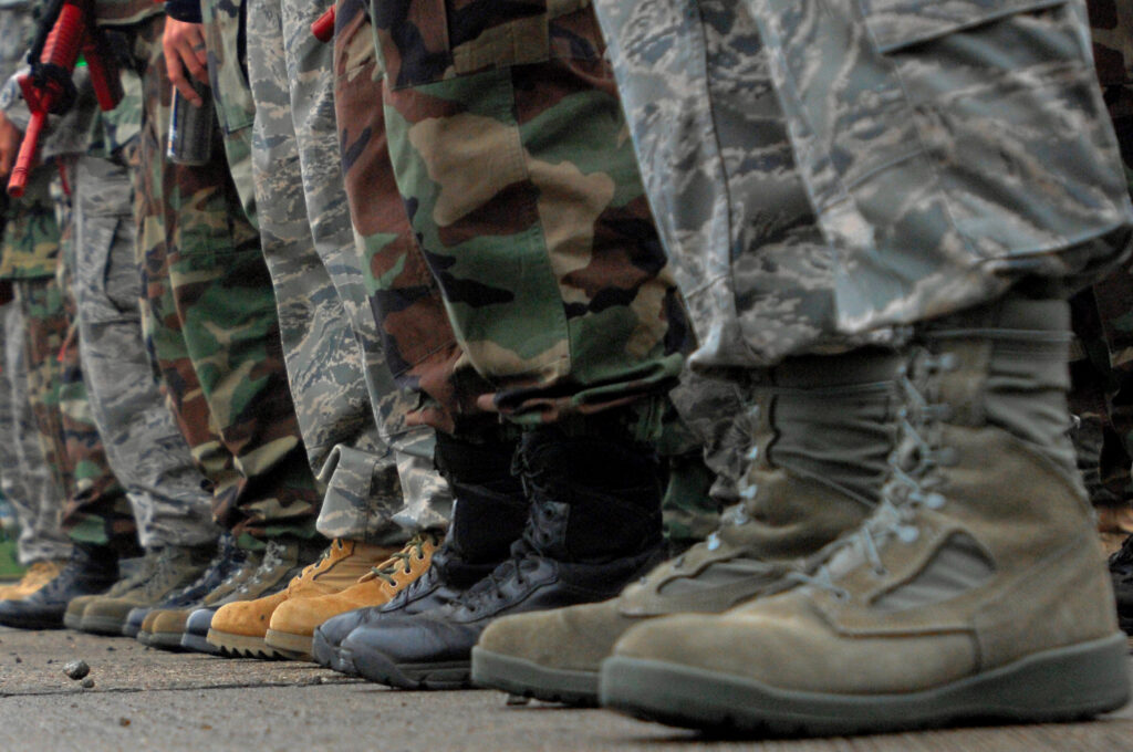 A close-up, ground-level view of a row of military personnel standing side by side, showing only their lower legs and boots. They are wearing various camouflage uniforms and tactical footwear, standing on a concrete surface.