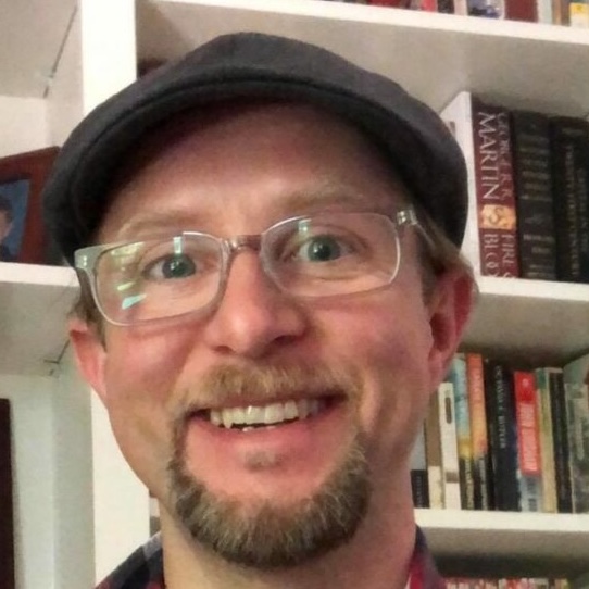 A smiling man wearing glasses and a dark flat cap, with a light mustache and goatee. He is standing in front of a white bookshelf filled with books.