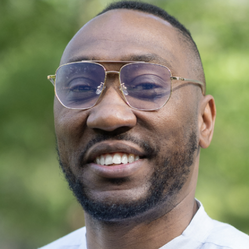 A man wearing gold-rimmed glasses and a white button-up shirt smiles at the camera. He has a neatly trimmed beard and short hair. The background is softly blurred with green foliage, suggesting an outdoor setting.