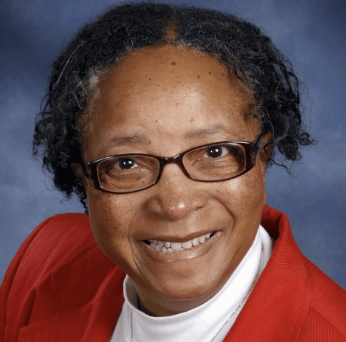Portrait of an older woman smiling warmly at the camera. She is wearing glasses, a white turtleneck, and a bright red blazer. Her hair is short and naturally curly, and the background is a soft blue studio backdrop.