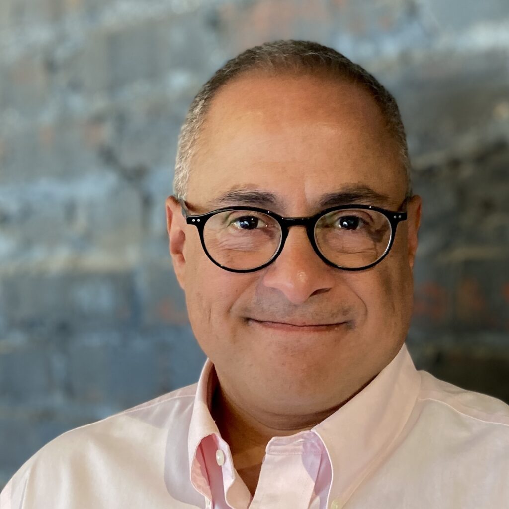 A man with short gray hair and black-rimmed glasses smiles softly at the camera. He is wearing a light pink button-down shirt and stands in front of a blurred gray brick wall.