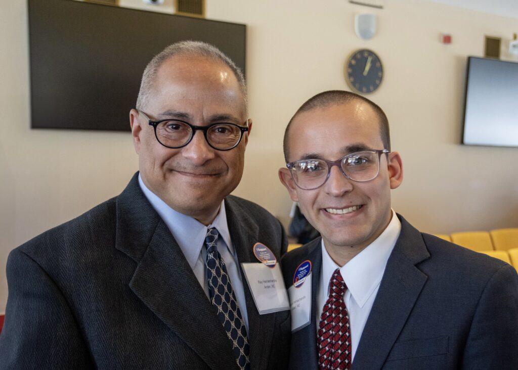 Two men in suits smile while posing for a photo indoors. Both are wearing glasses and name tags; one man has graying hair and a blue patterned tie, and the other has closely cropped hair and a red patterned tie. They each wear a round "I Support Public Education" button. Behind them are a clock, a television screen, and yellow chairs.
