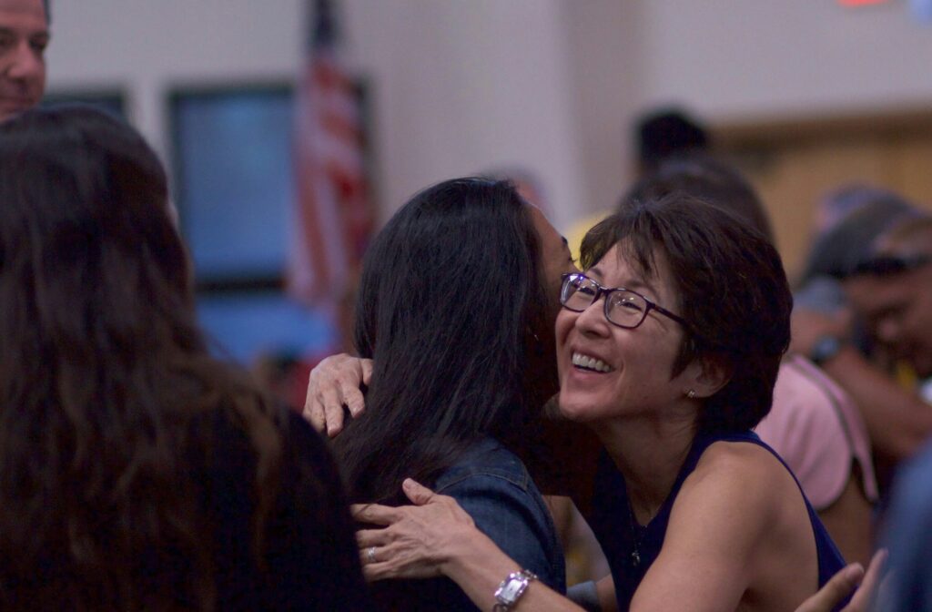A smiling woman wearing glasses warmly hugs another woman at an indoor gathering. The background is softly blurred, showing several people and a U.S. flag, suggesting a community or social event.
