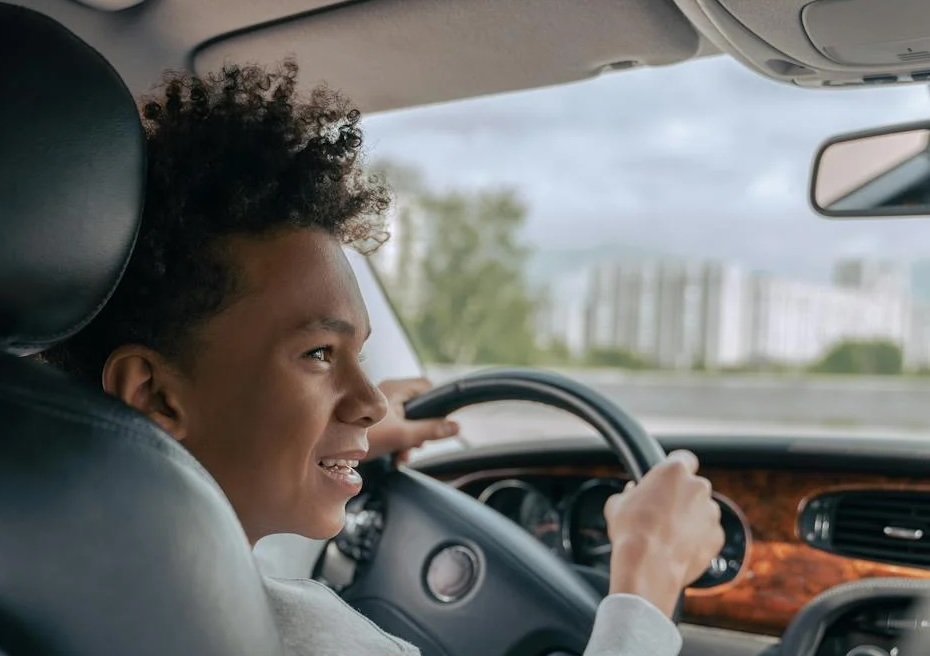 A smiling teenage boy driving a car, viewed from the back seat. He has curly hair and is gripping the steering wheel with both hands while looking at the road ahead. The interior of the car features a wood-paneled dashboard, and a cityscape is visible through the windshield.
