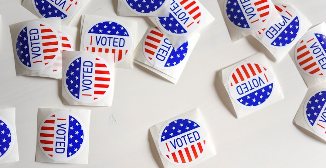 A scattered arrangement of round 'I Voted' stickers on a white surface. The stickers are red, white, and blue, with stars and stripes resembling the American flag.