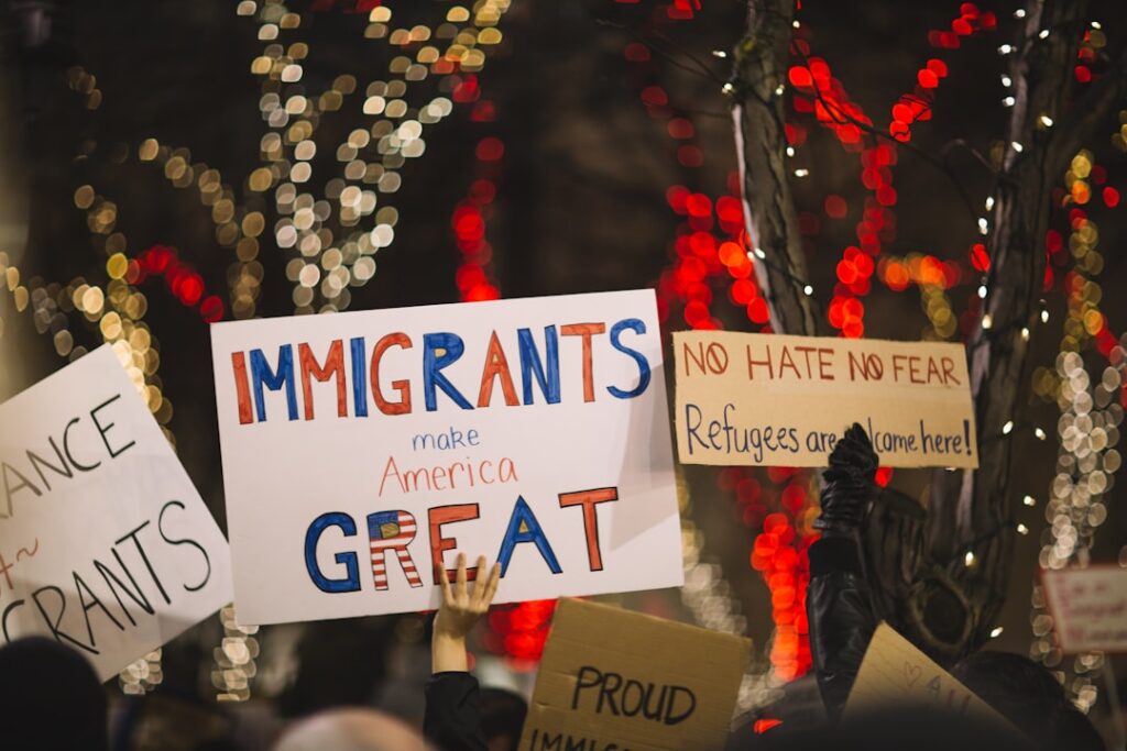 Protesters hold handmade signs supporting immigrants and refugees, including one that reads ‘Immigrants make America GREAT’ and another that says ‘No hate, no fear. Refugees are welcome here!’ The scene is set at night with trees decorated in red and white holiday lights.