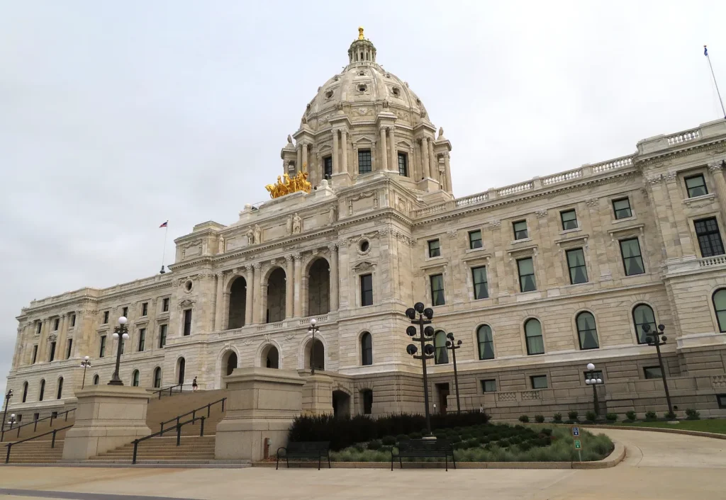 Exterior view of the Minnesota State Capitol building in St. Paul, featuring a large white marble facade with arched entryways, rows of rectangular windows, a central dome topped with a gold statue, and a golden quadriga sculpture at the base of the dome. The surrounding plaza includes staircases, benches, and ornamental street lamps under a cloudy sky.
