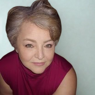 Woman with short, styled grayish-brown hair wearing a sleeveless burgundy top, looking up toward the camera with a soft smile and a light teal background.