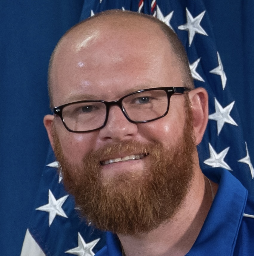 Smiling man with a reddish beard, glasses, and a blue collared shirt, standing in front of an American flag backdrop.