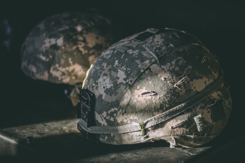 Close-up of two U.S. Army helmets with digital camouflage pattern, resting on a dark surface. The foreground helmet shows signs of wear and stitching repairs, with dramatic lighting highlighting its texture.
