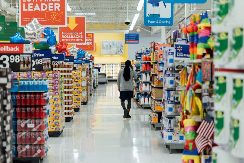 A person with long dark hair walks down the center aisle of a brightly lit Walmart store. Shelves on both sides are stocked with cleaning products, snacks, beverages, and piñatas. Overhead signs advertise low prices and rollback deals.