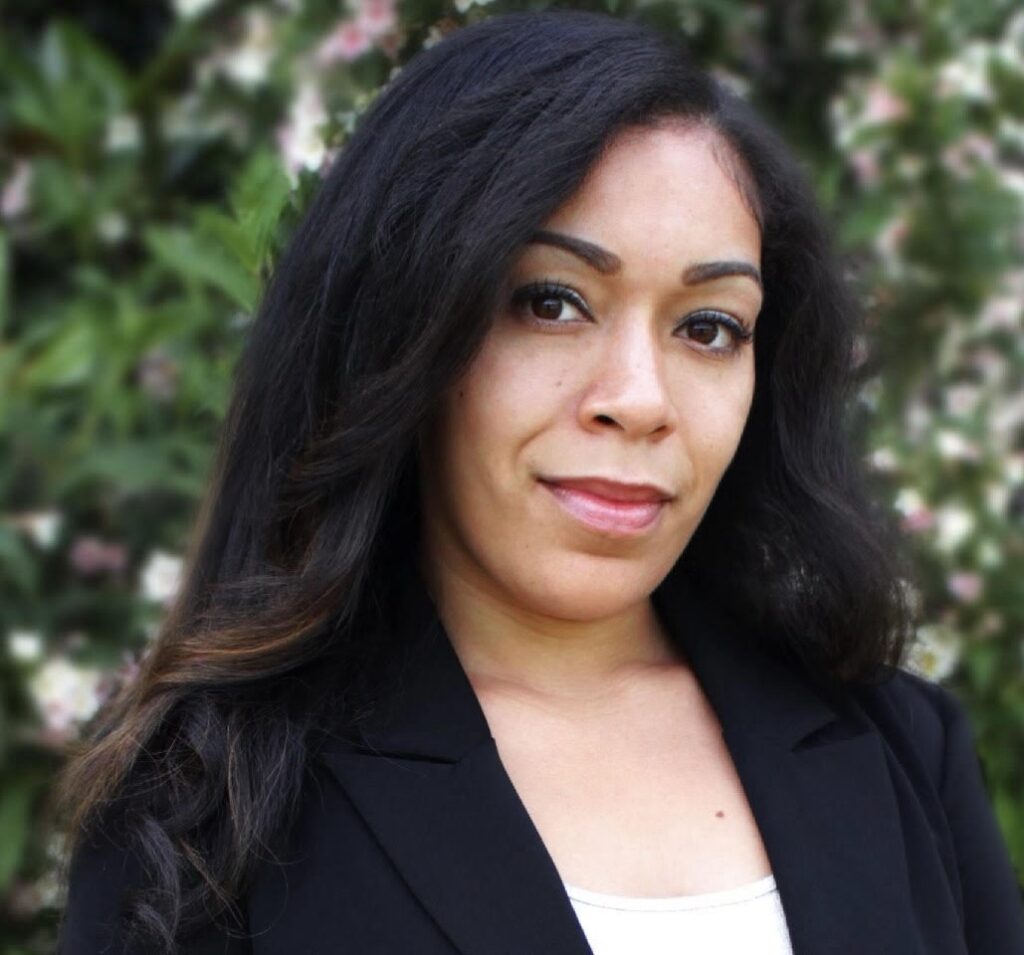 Crystal Barnes, a woman with long, dark hair, wearing a black blazer over a white top. She is standing outdoors in front of a blurred background of green foliage and light pink flowers, looking confidently at the camera with a slight smile.
