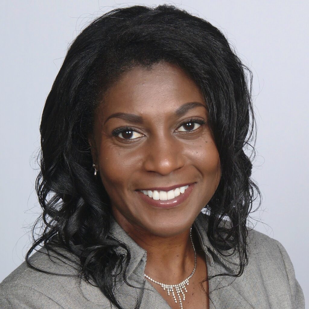 Rev. Della Owens, a woman with long, wavy black hair, smiles warmly at the camera. She is wearing a light gray blazer and a delicate, sparkling necklace, with a soft gray background behind her.