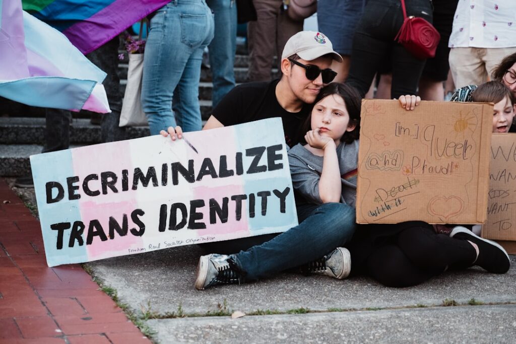 A group of people sitting on the ground at a protest or rally. One person holds a sign that reads “DECRIMINALIZE TRANS IDENTITY” with pastel pink, blue, and white colors resembling the transgender pride flag. A child next to them holds a cardboard sign that says “I’m queer and proud! Deal with it!” with colorful drawings. Other protest signs and pride flags are visible in the background.