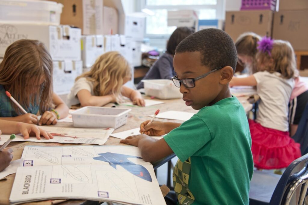 A classroom scene with young children sitting at a table and working on assignments. The children are focused, using pencils and looking at papers or books. One child in the foreground wears a green shirt and glasses, intently reading and writing. The classroom is filled with educational materials and storage boxes. Natural light comes in from a window in the background.