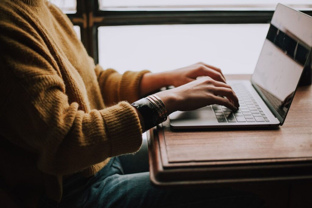 A person wearing a mustard yellow sweater and blue jeans is typing on a laptop at a wooden desk. Only their torso and hands are visible. The background shows a window with natural light coming through, creating a cozy and focused atmosphere.