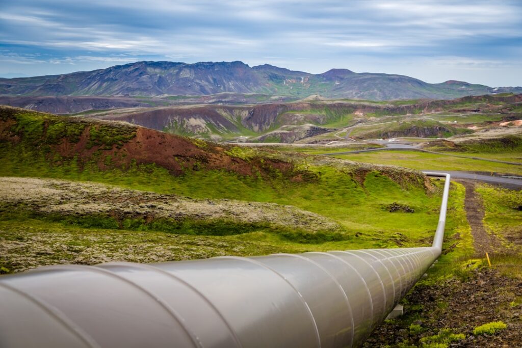 A large industrial pipeline stretches across a vibrant green landscape with rolling hills and rocky terrain. In the background, there are distant mountains under a cloudy blue sky. The pipeline appears to be part of an energy or utility infrastructure, cutting through the natural scenery.