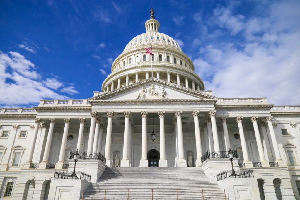 The United States Capitol building in Washington, D.C., viewed from the front. The neoclassical white structure features a large central dome topped with the Statue of Freedom, rows of columns, and a wide staircase leading up to the entrance. An American flag flies above the portico under a partly cloudy blue sky.